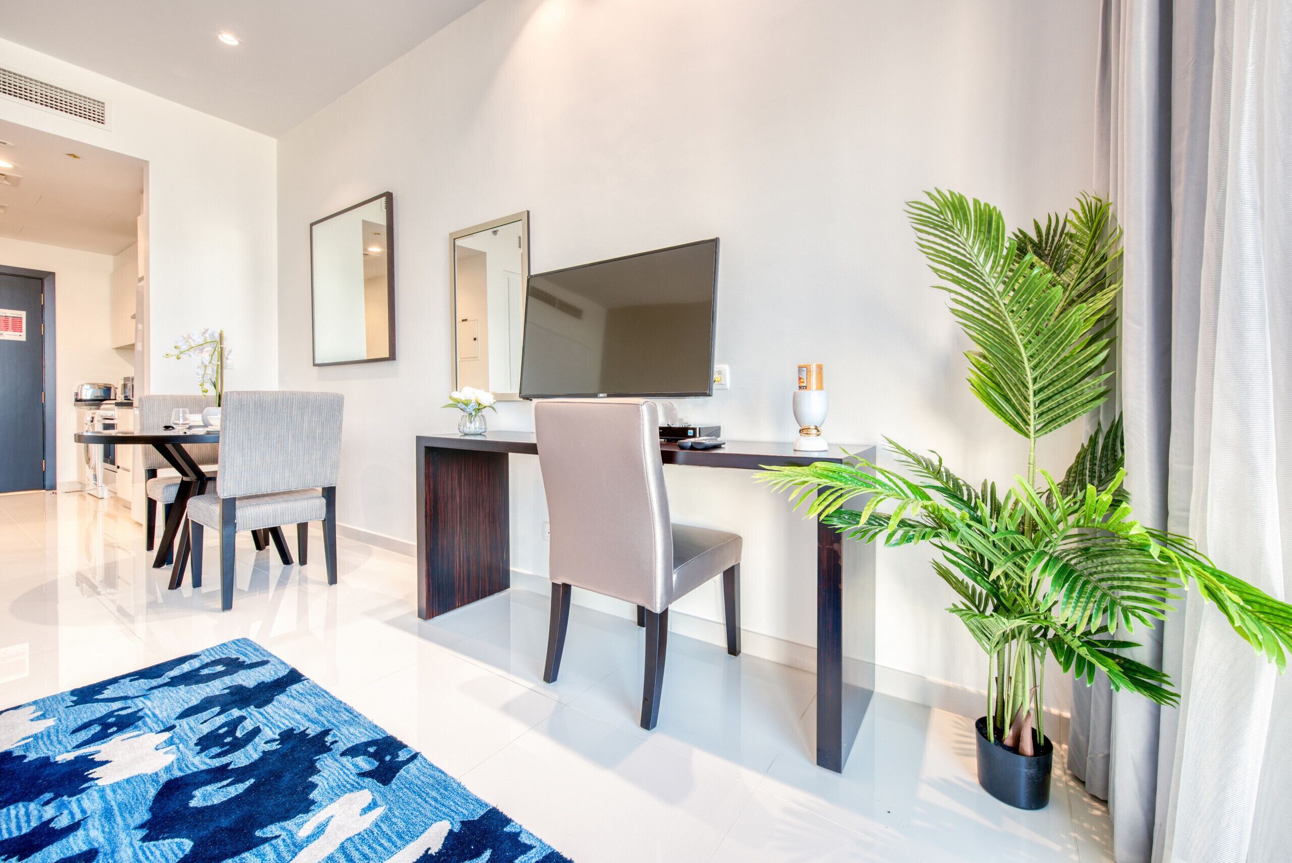 A modern hotel room with a dining area, a large flat-screen TV, a potted plant, and a blue patterned rug.