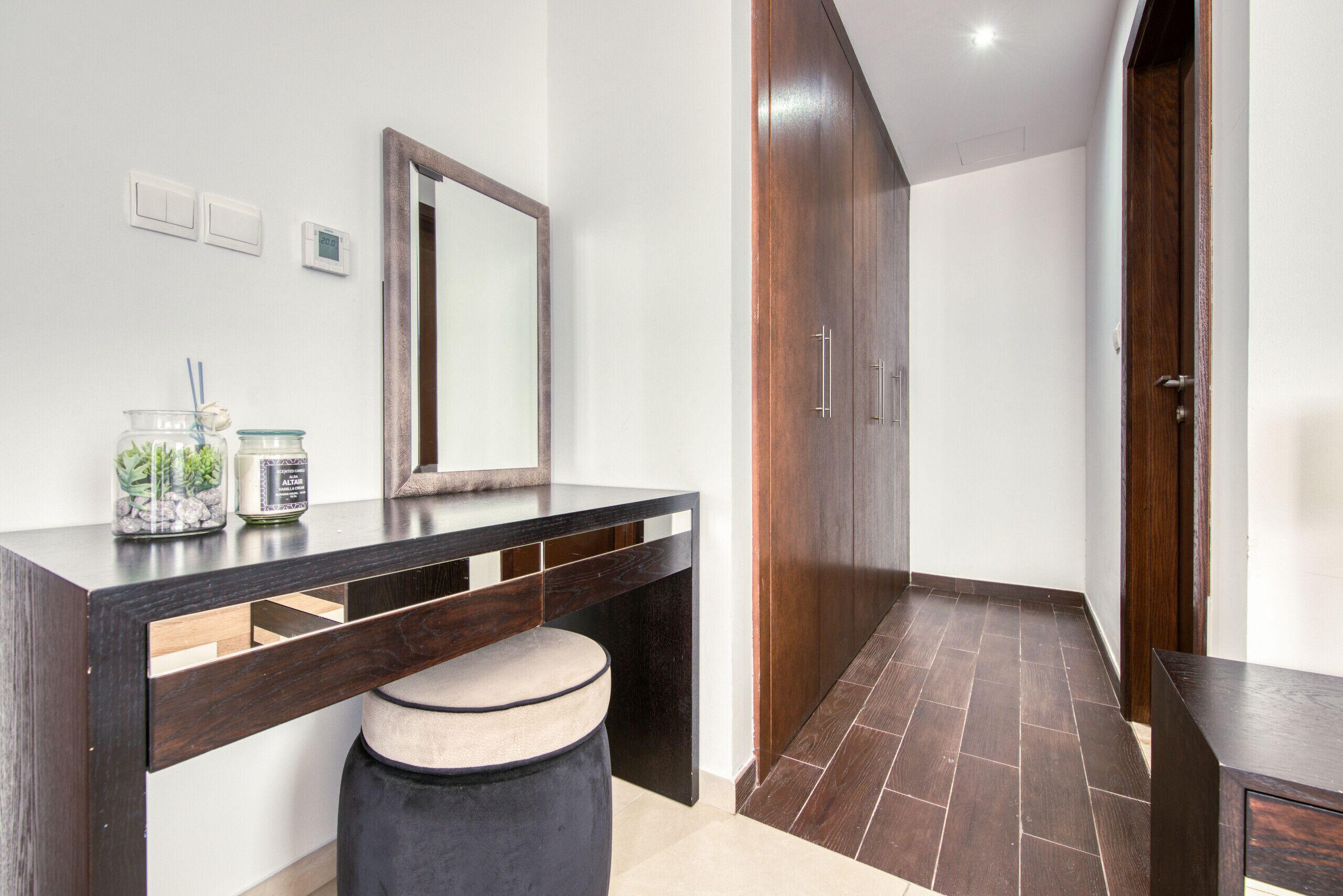 A modern bathroom with a dark wooden vanity, a round stool, a mirror, and a shower area with dark tiles.