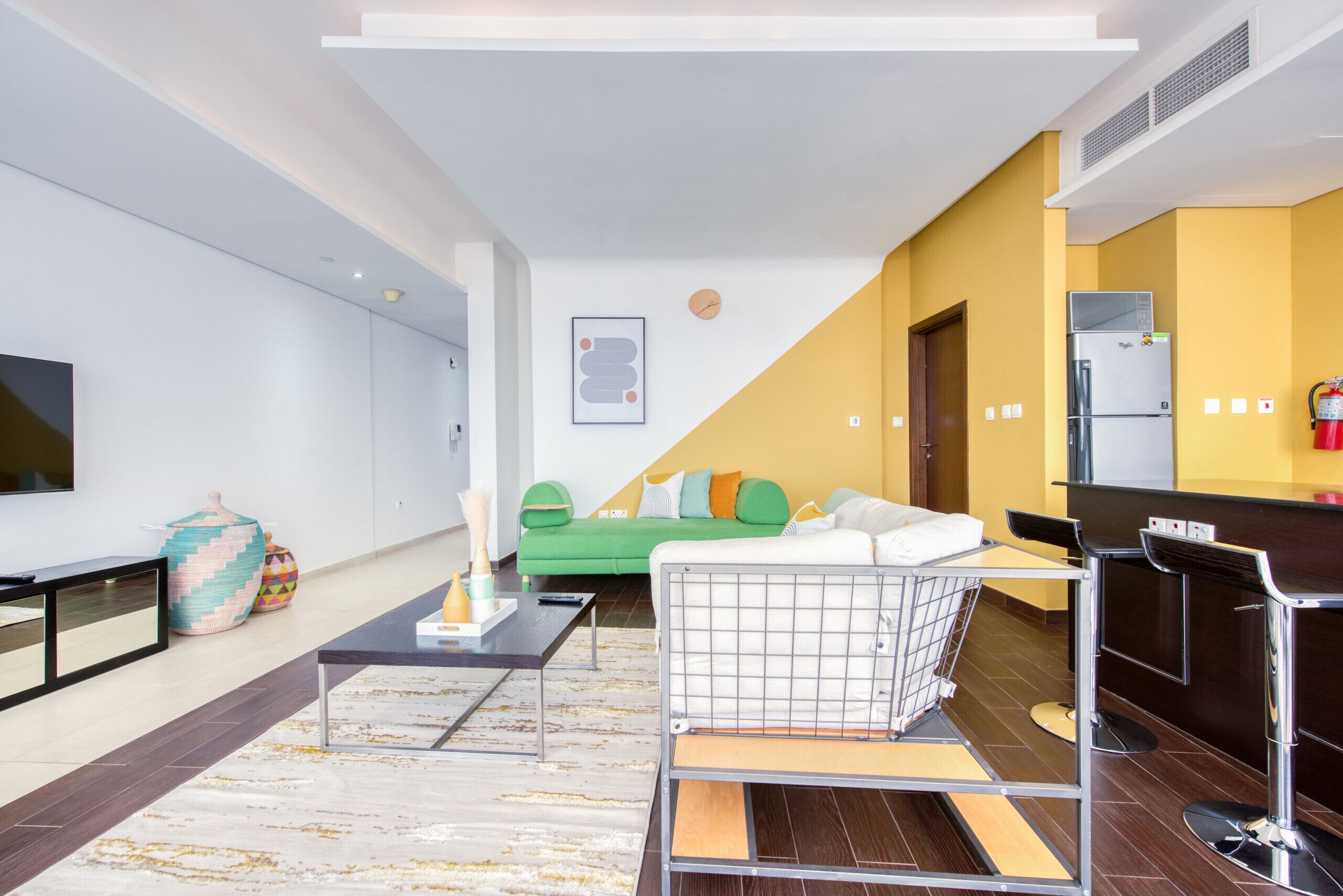 A modern living room with a green sofa, a coffee table, and a kitchen area with a refrigerator.