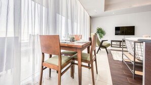 Uma área de refeições moderna com mesa e cadeiras de madeira, uma televisão na parede e uma cama com estrutura de metal ao fundo.