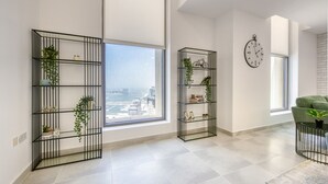 A modern living room with large windows, black metal shelving units with plants, a green sofa, and a wall clock. - Outstanding 3BR at Sadaf 7 JBR by Deluxe Holiday Homes (Dubai)