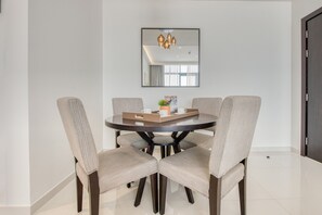Uma sala de jantar com uma mesa redonda, quatro cadeiras estofadas, uma bandeja com chávenas e uma planta, além de um candelabro acima.