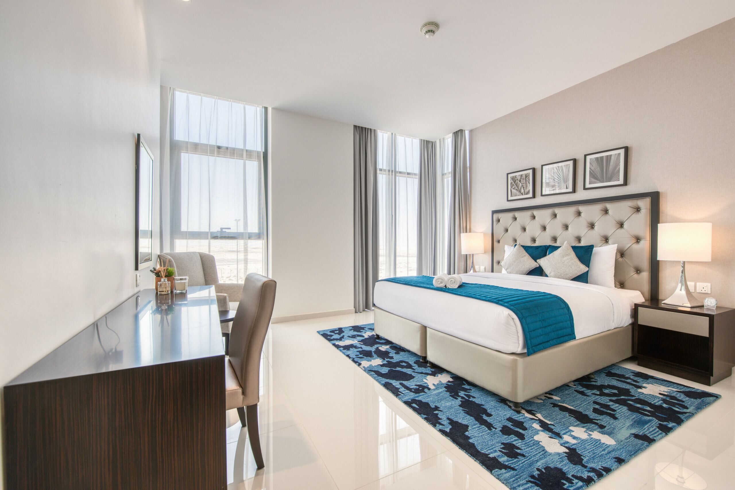A modern hotel room with a large bed, a desk, and a chair. There are large windows with curtains, a headboard with a tufted design, and a blue patterned rug.
