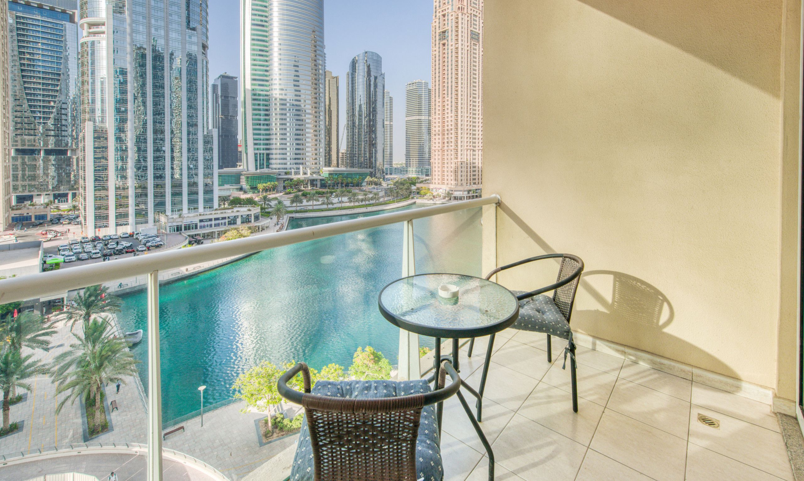 A balcony with a glass table and wicker chairs overlooking a marina with tall buildings in the background.