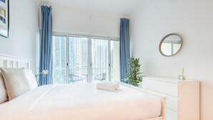 A hotel room with a large bed, a round mirror, a dresser, and a view of tall buildings through the window.
