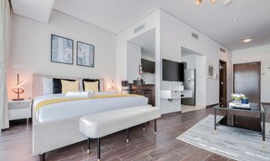 A modern hotel room with a large bed, bedside tables, a bench, a TV, a kitchen area, and a bathroom. - Cozy Studio at The Matrix Sports City by Deluxe Holiday Homes (Dubai)