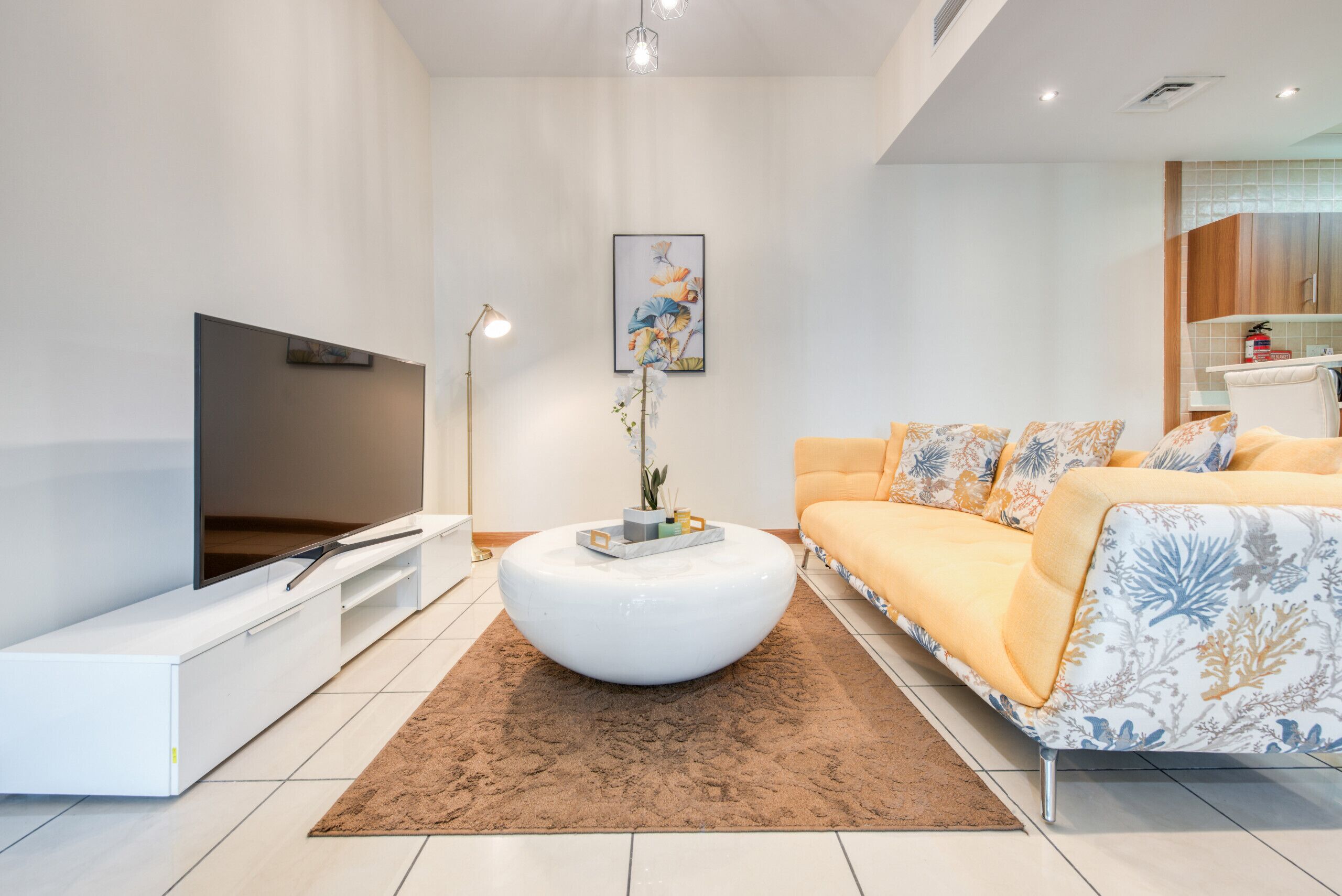A modern living room with a flat-screen TV, a white coffee table, a sofa with floral cushions, and a framed artwork on the wall.