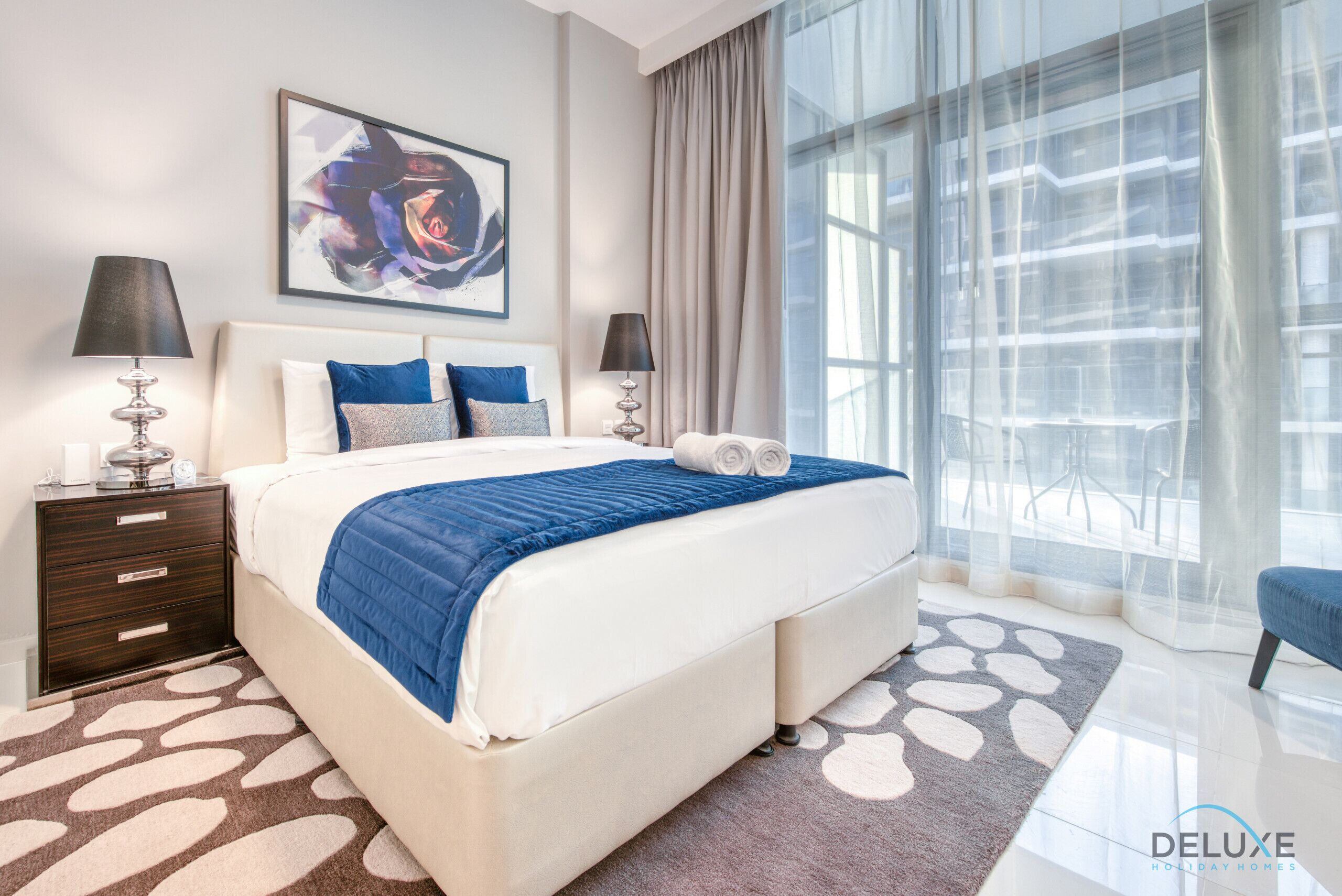 A modern hotel room with a large bed, two bedside tables, a nightstand, a large window with sheer curtains, and a decorative rug.