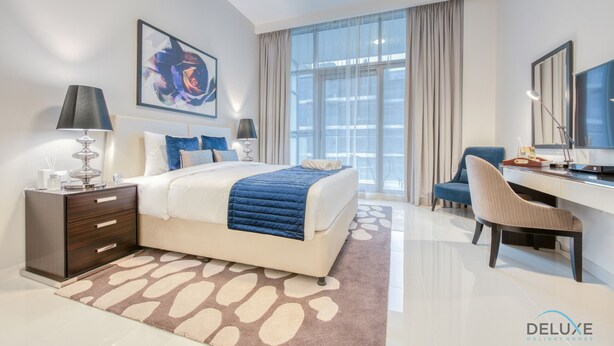 A modern hotel room with a large bed, a desk, and a chair. There is a window with curtains, a painting on the wall, and a logo on the floor.