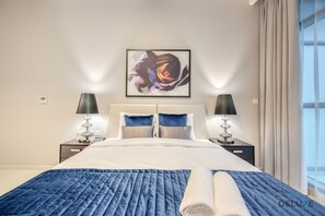 A hotel room with a large bed, two bedside tables with lamps, a painting on the wall, and a view of the outdoors. - Stunning Studio at Golf Promenade 2A Damac Hills by Deluxe Holiday Homes (Dubai)