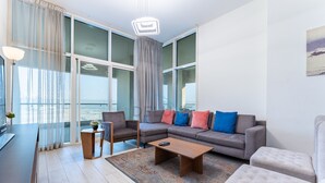 A modern living room with a grey sofa, a wooden coffee table, and a grey armchair. There is a large window with curtains, a view of the city, and a painting on the wall.