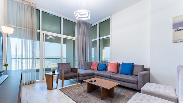 A modern living room with a grey sofa, a wooden coffee table, and a grey armchair. There is a large window with curtains, a view of the city, and a painting on the wall. - Huge Balcony Overlooking Creek Harbour with Glimpses of Burj Khalifa (Dubai)