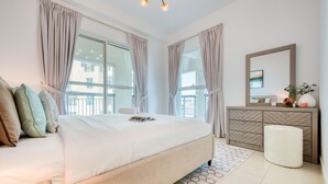 A modern bedroom with a large bed, a dresser, a mirror, and a view of the outside balcony.