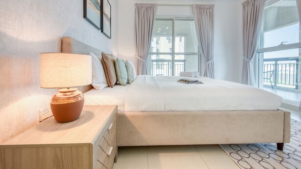 A hotel room with a large bed, a bedside table with a lamp, a window with curtains, and a view of the outside balcony.