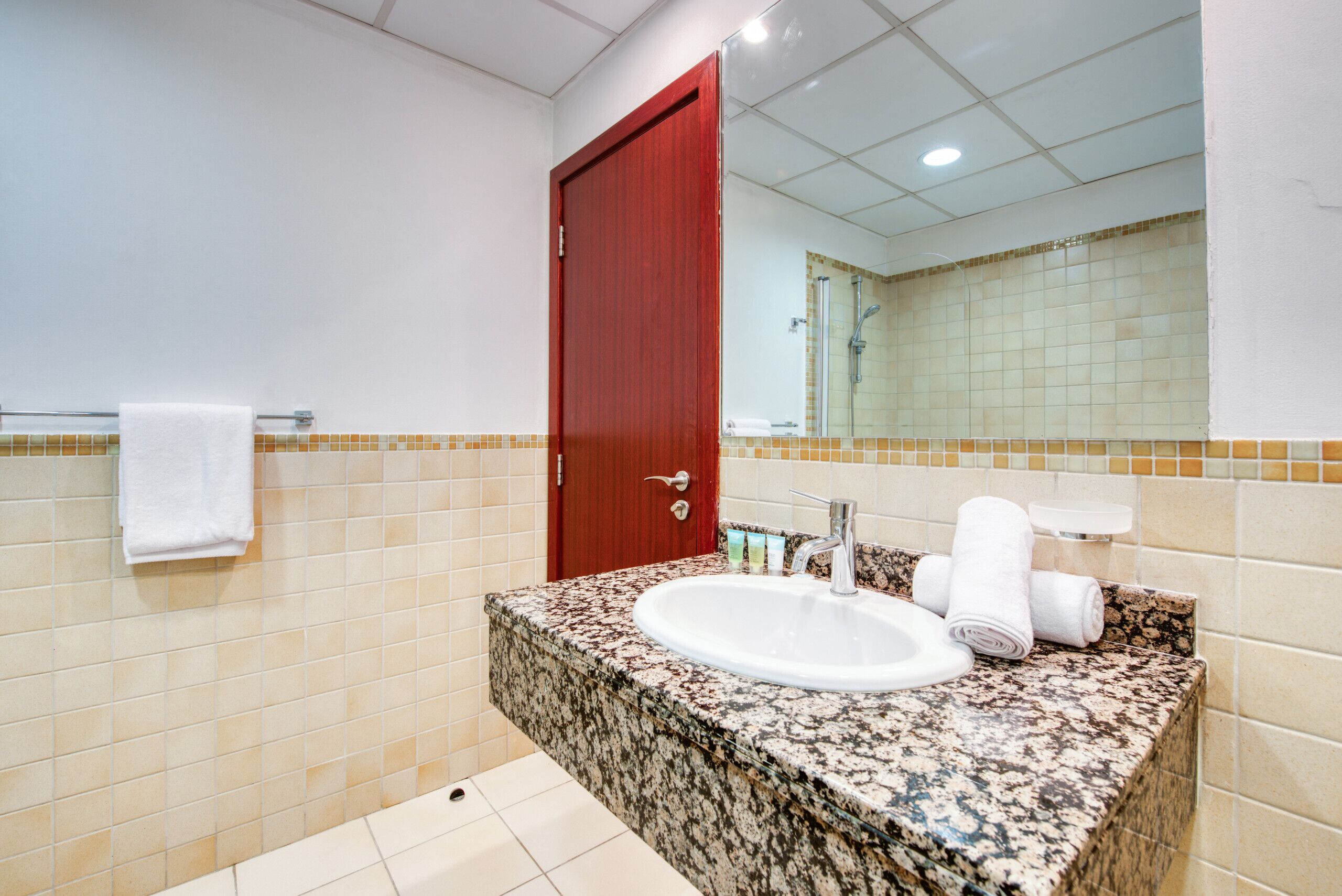 Une salle de bain avec un comptoir en granit, un lavabo blanc, une porte rouge et un miroir qui reflète l’espace de la douche.