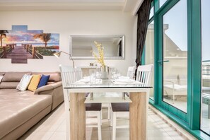 A modern living room with a glass dining table, a beige sofa, and a large window with a view of the city.