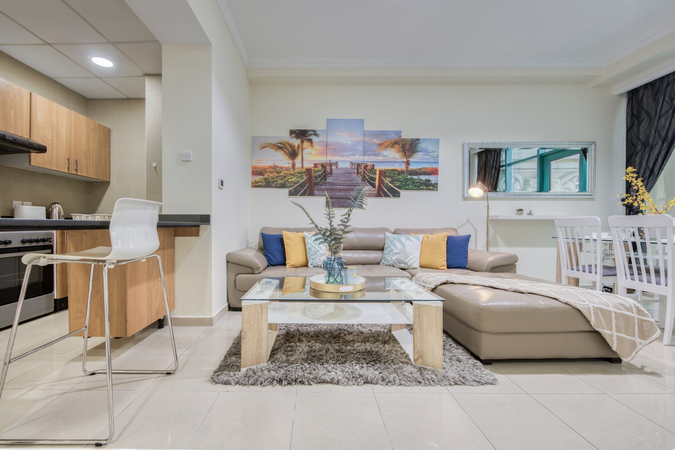 A modern living room with a glass coffee table, a sofa, and a dining area with a white chair. There is a kitchen with wooden cabinets and a stove.