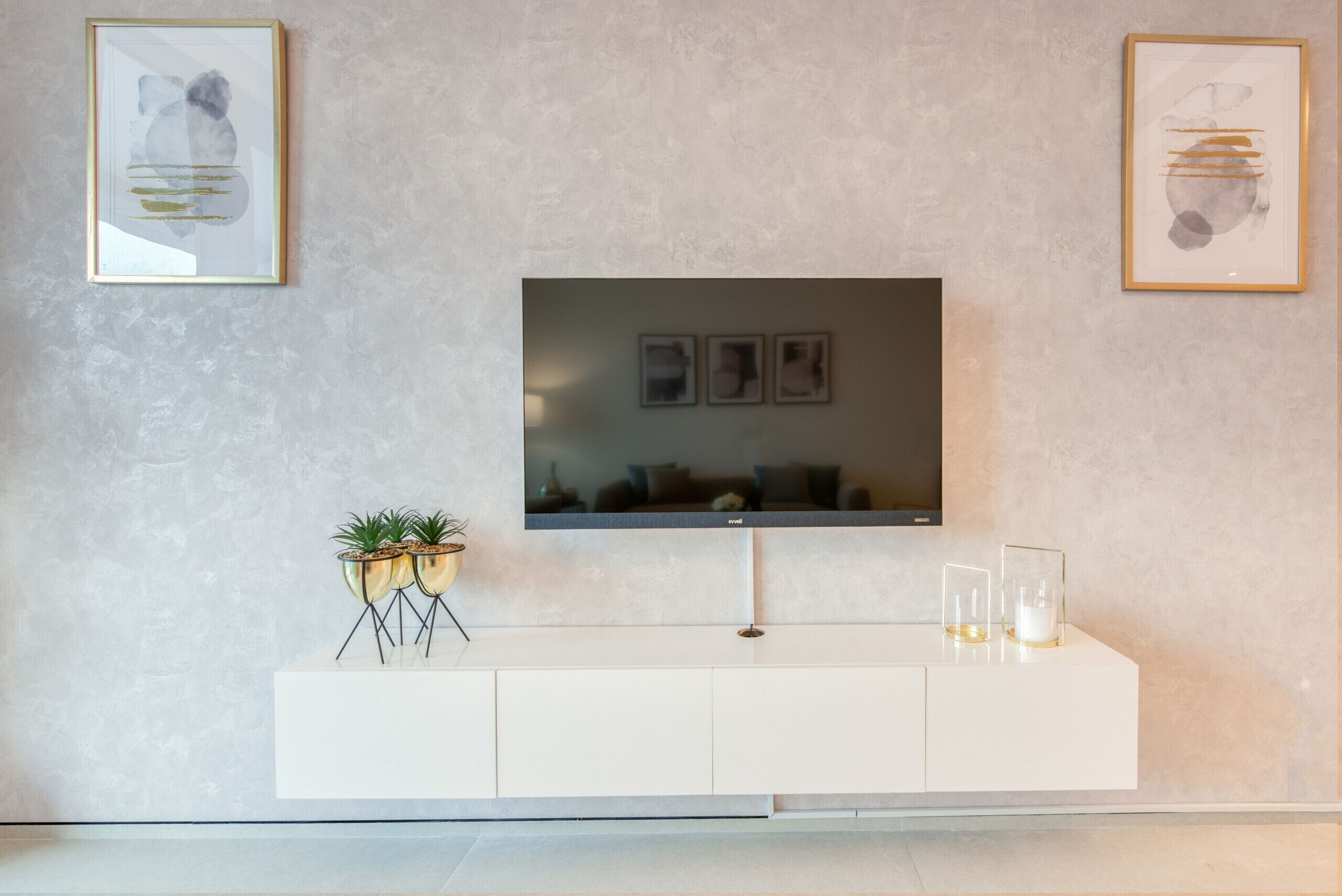 Ein modernes Wohnzimmer mit einem flachbildfernseher, der an einem weißen Schrank befestigt ist, begleitet von zwei gerahmten abstrakten Gemälden und einer kleinen Pflanzengruppe.