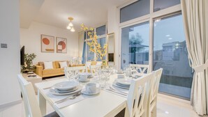 Ein Essbereich mit einem weißen Tisch, gedeckt für vier Personen, umgeben von weißen Stühlen. Ein großes Fenster mit Vorhängen, eine Couch im Hintergrund und ein Kunstwerk an der Wand.