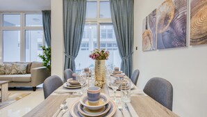 A dining area with a wooden table set for six, a vase with flowers, and a view of the city through large windows.