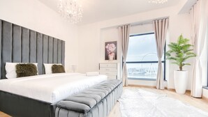 A modern bedroom with a large bed, a grey upholstered bench, a white rug, a potted plant, and a view of a bridge through the window. - Elegant 2BR at Bahar 2 JBR by Deluxe Holiday Homes (Dubai)