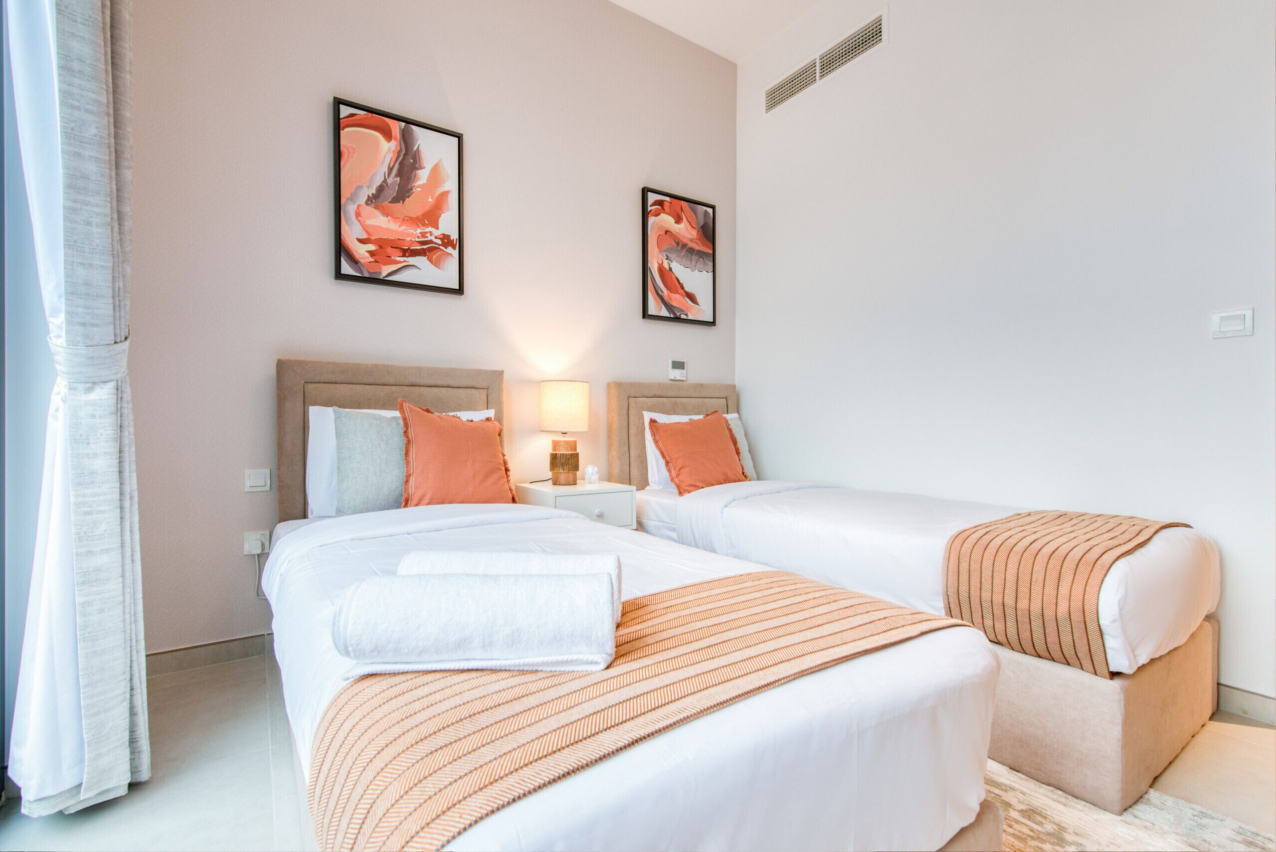 Een hotelkamer met twee bedden, elk met een wit-oranje gestreept dekbed, een nachtkastje met een lamp en twee ingelijste abstracte schilderijen aan de muur.