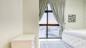 Une chambre avec une grande fenêtre, un lit doté d’une tête de lit capitonnée, une commode blanche et un tapis bleu à motifs.