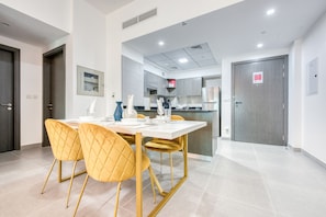 Uma sala de jantar moderna com uma mesa branca e cadeiras douradas, uma cozinha ao fundo e uma porta fechada com uma placa.