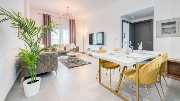 A modern living room with a sofa, coffee table, and a dining area with a table set for four. There is a large window with curtains, a flat-screen TV mounted on the wall, and a potted plant. - Whimsical 2BR at Bella Rose Barsha South by Deluxe Holiday Homes (Dubai)
