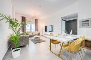 Uma sala de estar moderna com sofá, mesa de centro e área de jantar com mesa posta para quatro pessoas. Há uma janela grande com cortinas, uma TV de tela plana na parede e um vaso com planta.
