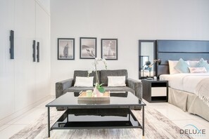 A modern bedroom with a grey sofa, a bed with a blue headboard, and a nightstand with a lamp. There are three framed pictures on the wall.