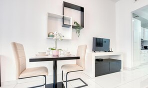 A modern dining area with a black table, two beige chairs, a small potted plant, and a television on a white cabinet.