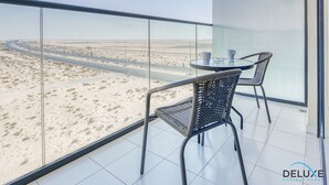 Een balkon met glazen hekwerk met uitzicht op een woestijnlandschap met een weg. Er staan twee rieten stoelen en een klein tafeltje met een kop en schotel.
