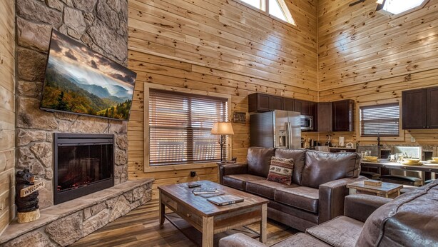 L’intérieur chaleureux d’un chalet avec une cheminée en pierre, des murs en bois, un canapé en cuir et un coin cuisine.