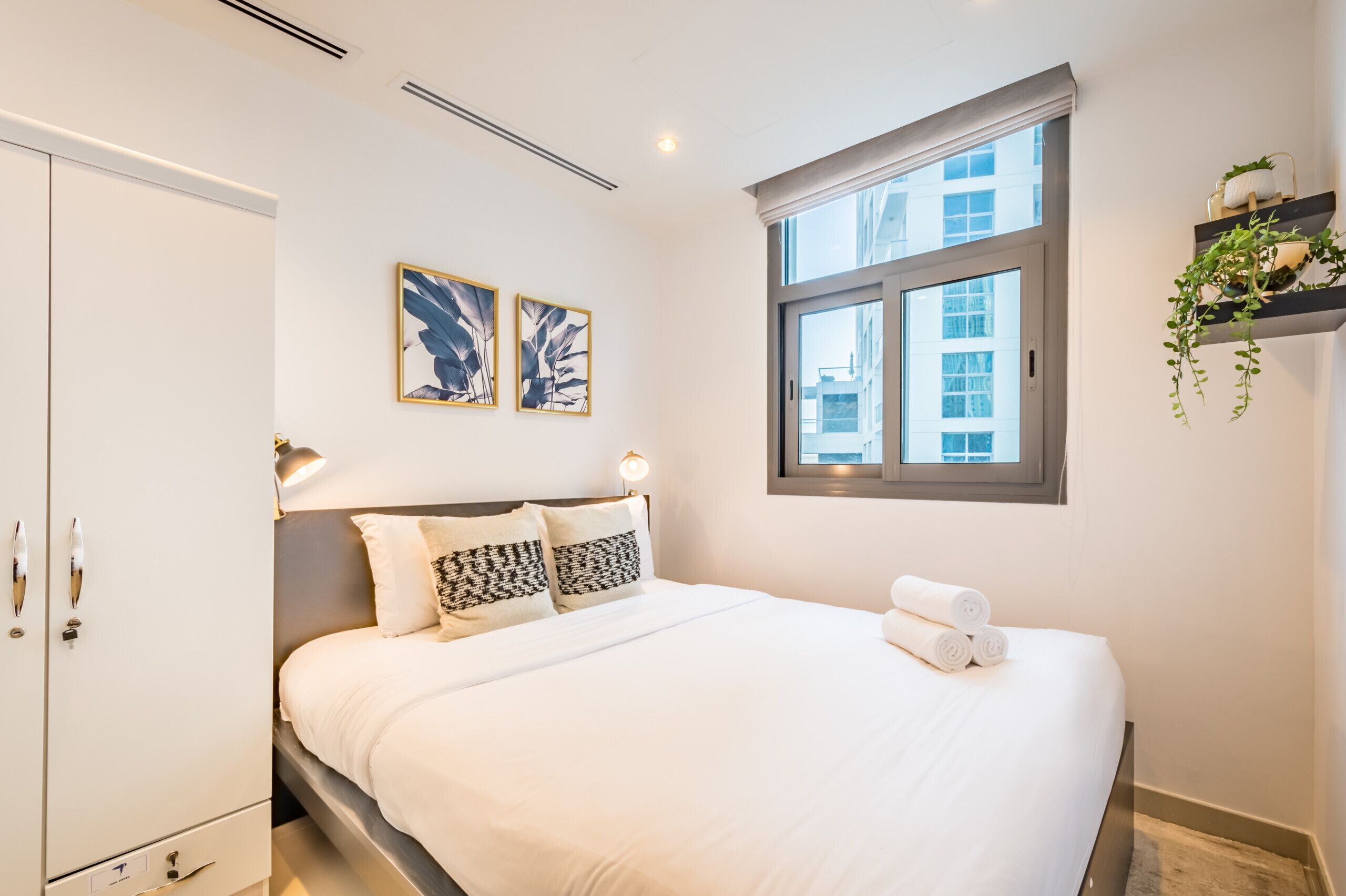 Habitación de hotel con una cama grande, un armario, dos obras de arte colgadas en la pared, una ventana con vistas a edificios y una planta en un estante.