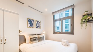 Une chambre d’hôtel avec un grand lit, une armoire, deux œuvres d’art accrochées au mur, une fenêtre donnant sur des immeubles et une plante posée sur une étagère.