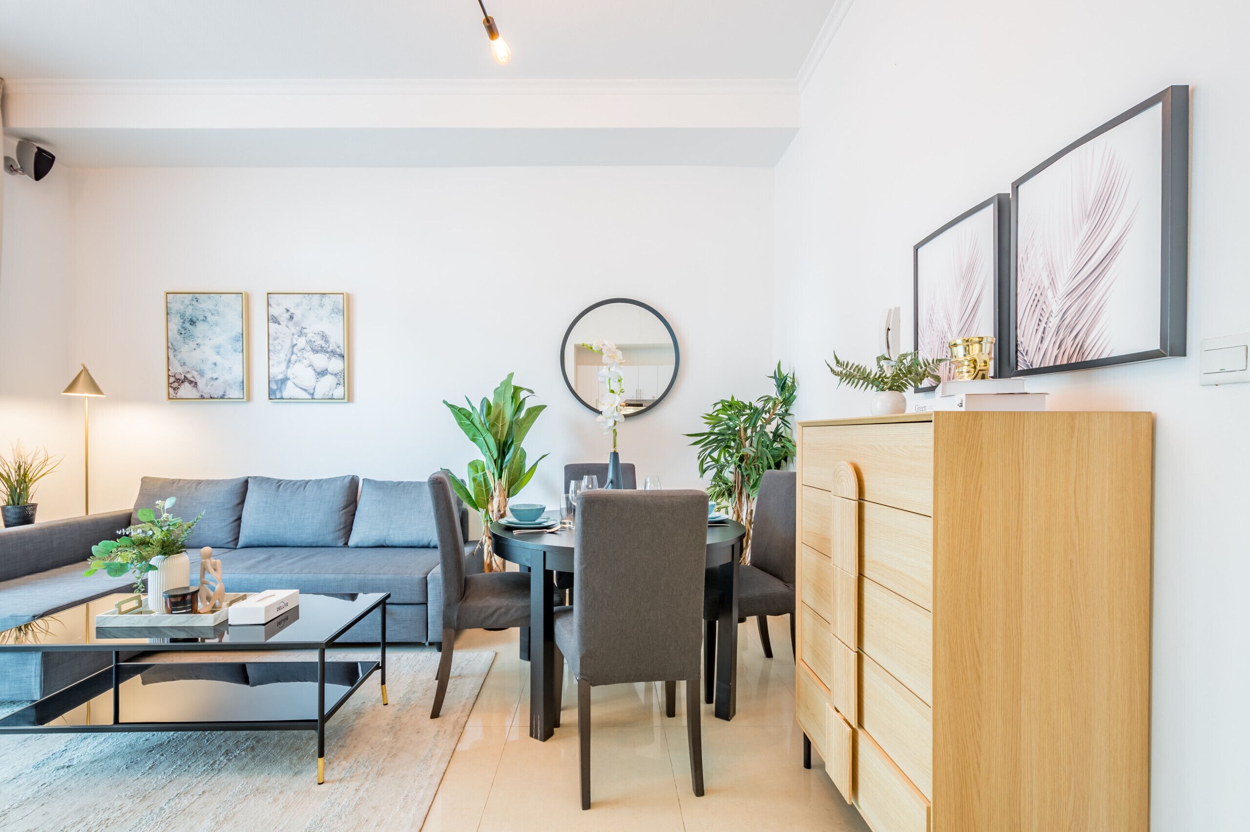 Een moderne woonkamer met een grijze bank, een salontafel, een eettafel met stoelen en een houten kast. Twee ingelijste kunstwerken hangen aan de muur en boven de eettafel hangt een ronde spiegel.