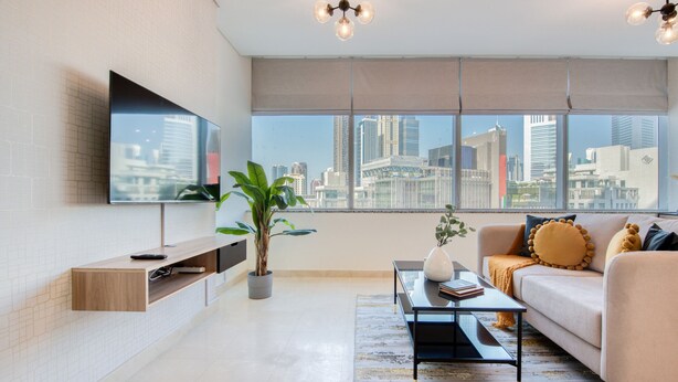 Una sala moderna con vista a la ciudad, un televisor de pantalla plana montado en la pared, un sofá con almohadones decorativos, una mesa de centro y una planta en maceta.