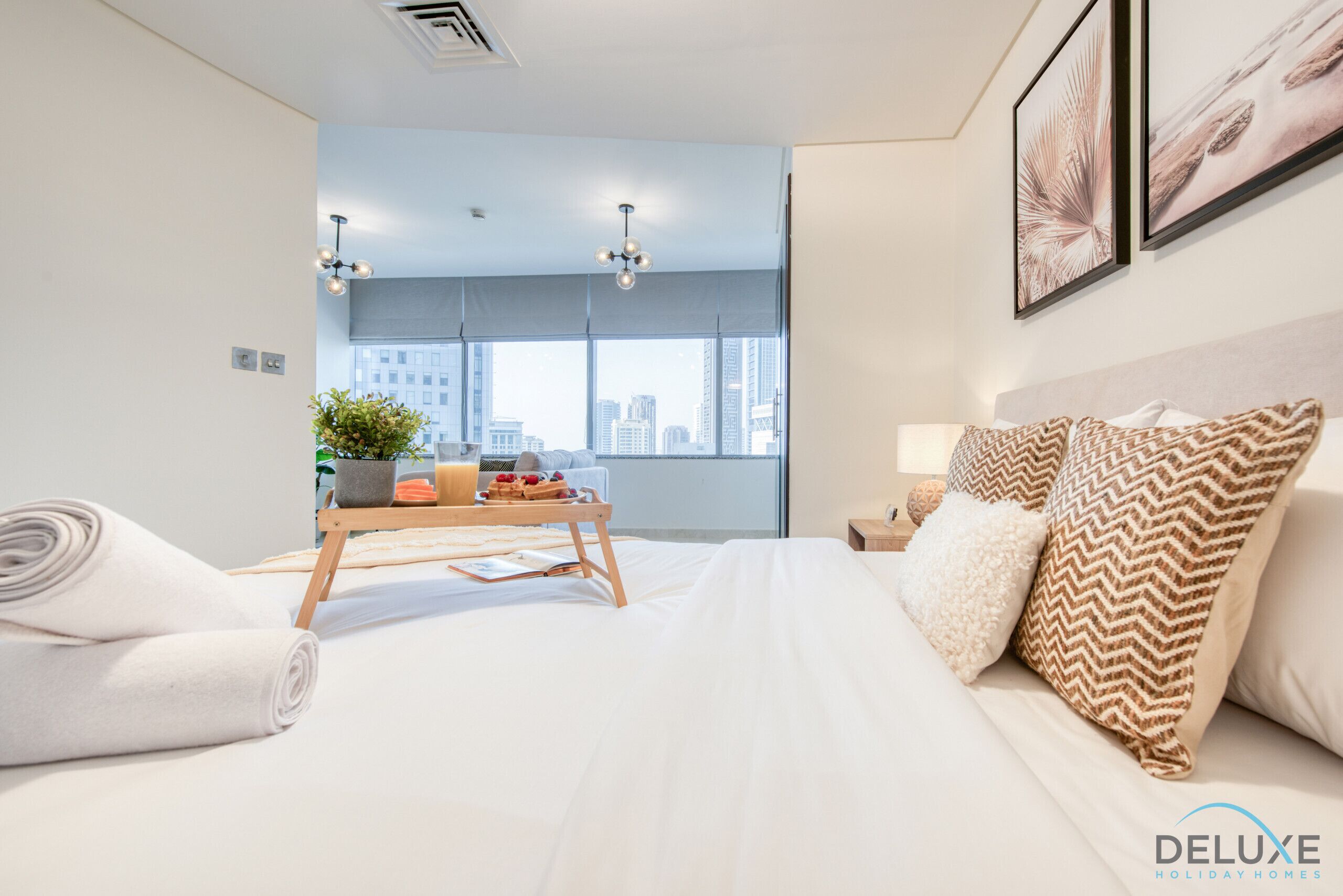 A modern hotel room with a large bed, a bedside table with a plant, and a view of the city through large windows.