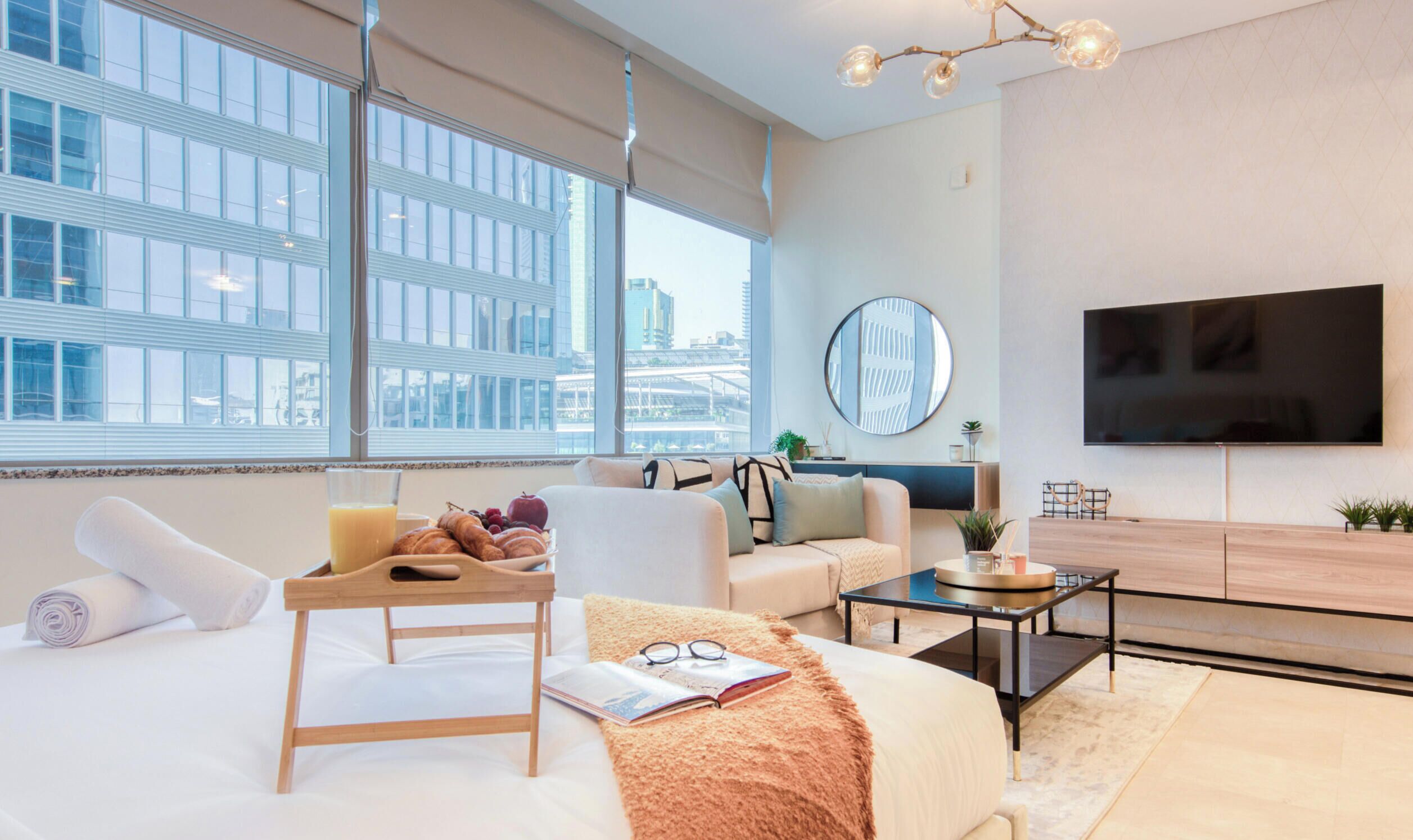 A modern hotel room with a large bed, a sitting area with a sofa and armchair, a dining table with breakfast items, a flat-screen TV, and a city view through large windows.