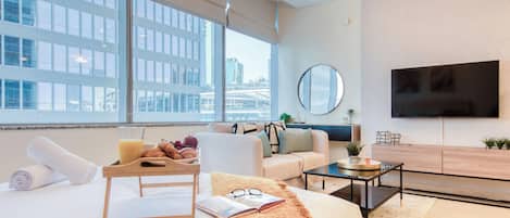 Habitación de hotel moderna con una cama grande, una zona de estar con sofá y sillón, una mesa de comedor con alimentos para el desayuno, un televisor de pantalla plana y vistas a la ciudad a través de amplios ventanales.