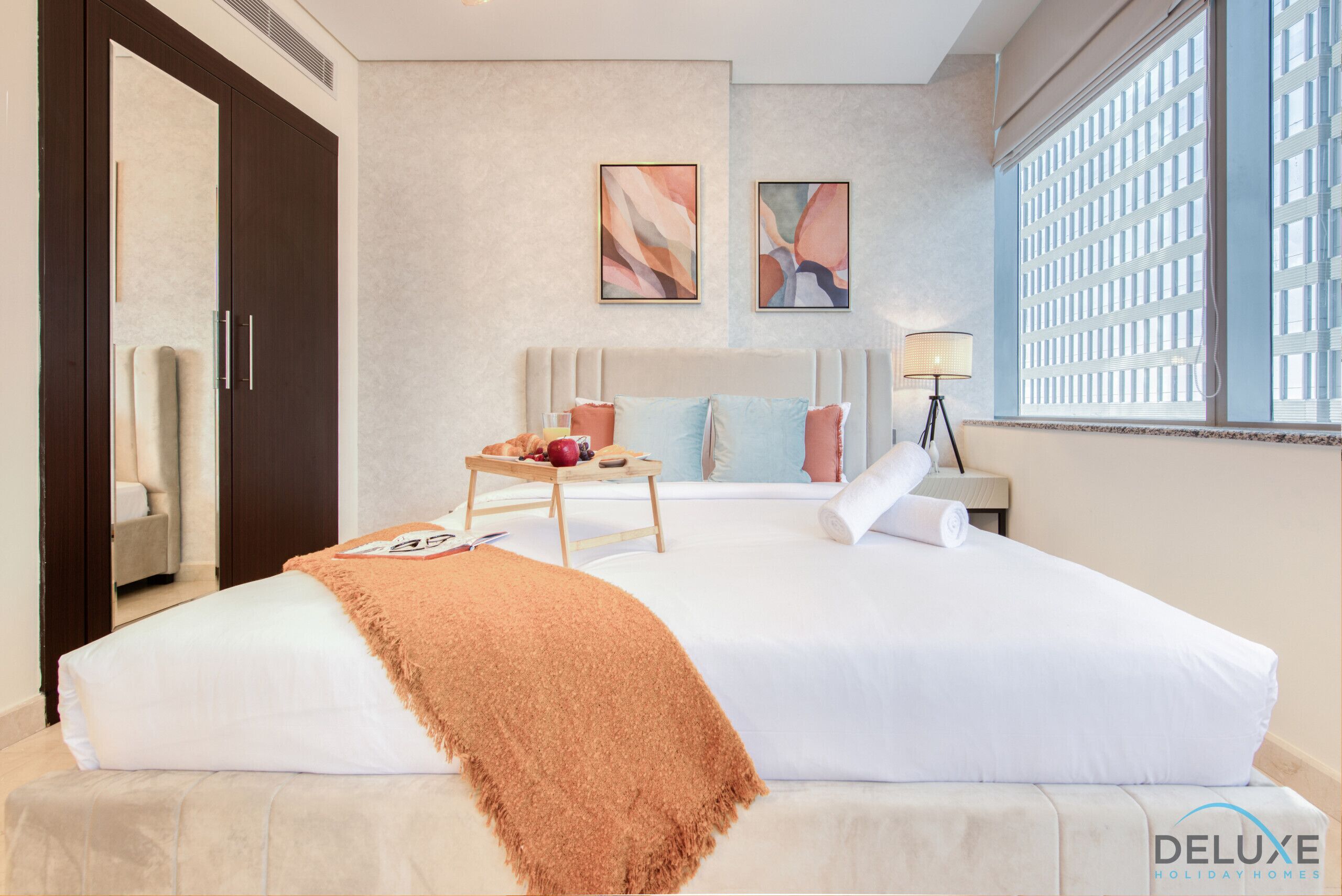 A hotel room with a bed, a bedside table with a lamp, a window with blinds, and artwork on the wall.
