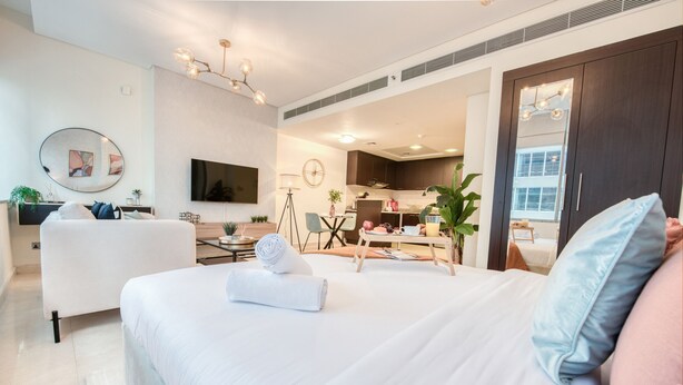 A modern hotel room with a large bed, a desk, and a kitchenette in the background.