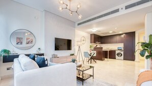 A modern living room with a sofa, coffee table, and a TV mounted on the wall. There is a kitchen area visible in the background with cabinets and a washing machine.