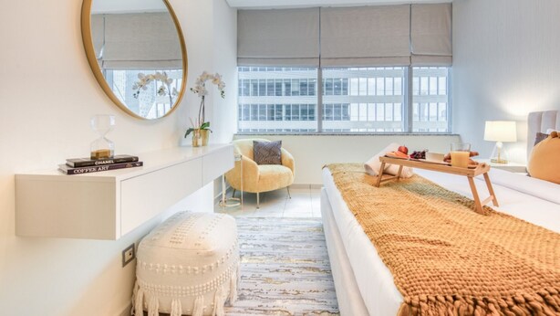 A modern bedroom with a large bed, a bedside table, a chair, a desk, a mirror, and a window with blinds. - Gorgeous 1BR at Sky Gardens DIFC by Deluxe Holiday Homes (Dubai)