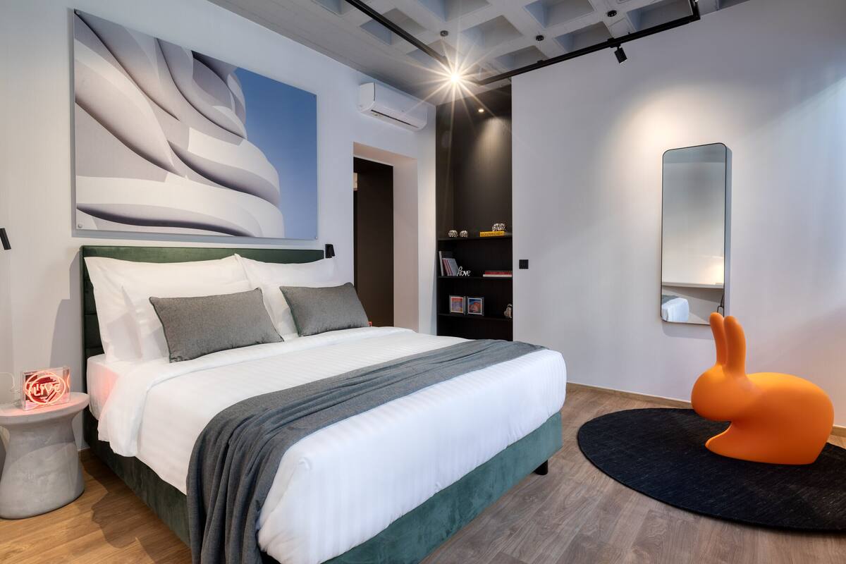 A modern bedroom with a large bed, a unique wall art piece, a small side table with a neon sign, and a distinctive orange rabbit sculpture.