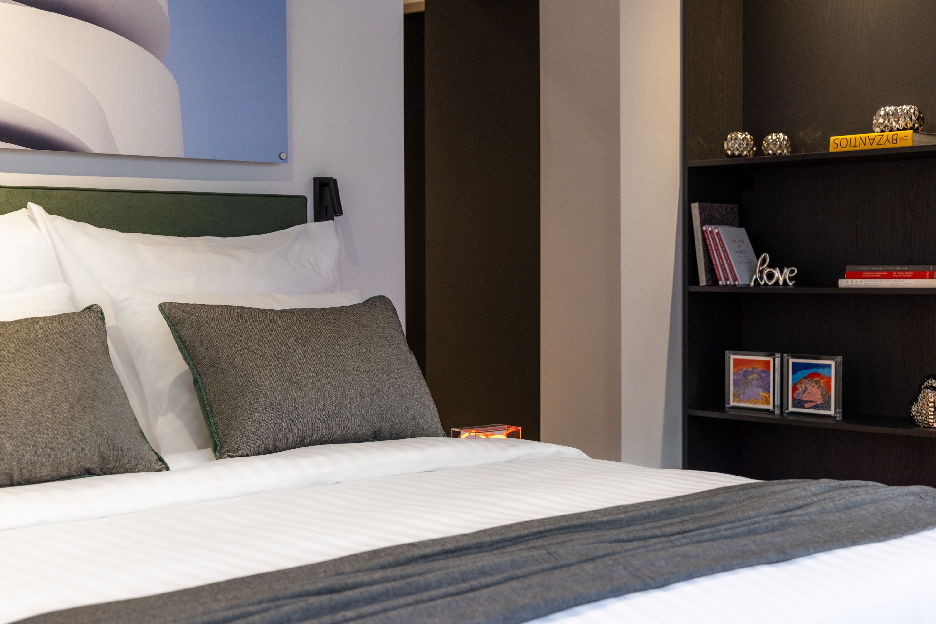 Una cama bien hecha con almohadas, una mesita de noche con un libro y un objeto decorativo pequeño, y una estantería con marcos y un libro titulado 'Love'.