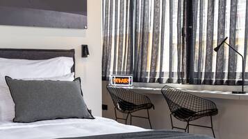 Une chambre d’hôtel moderne avec un lit, deux chaises en fil de fer noir, une fenêtre avec des stores verticaux et un panneau lumineux néon indiquant le mot « HÔTEL ».