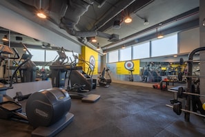 Uma academia com diversos equipamentos de exercícios, incluindo esteiras e pesos, e uma grande janela que proporciona iluminação natural.