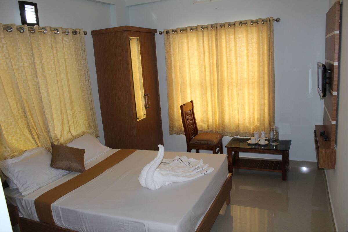 A hotel room with a bed, a wooden wardrobe, a desk with a chair, and a window with curtains.